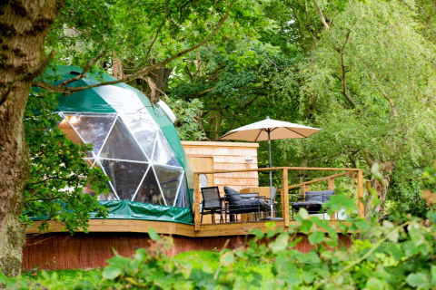 Chartwell Luxury Dome in the UK, nestled in lush green forest, featuring a wooden deck with outdoor seating.