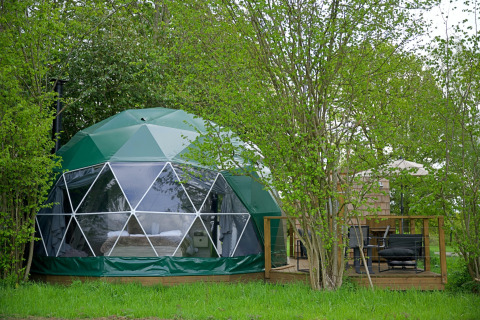 Hever Luxury Dome gemt blandt grønne træer med stor glasfacade og terrasse med havemøbler ved siden af.