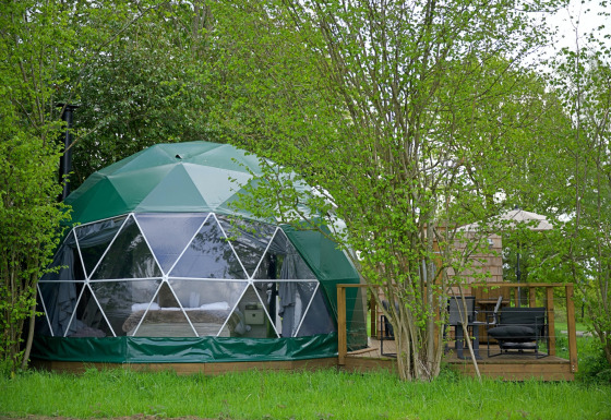 Hever Luxury Dome gemt blandt grønne træer med stor glasfacade og terrasse med havemøbler ved siden af.