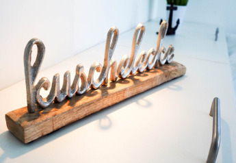 Escultura metálica con la palabra 'kuschelecke' sobre base de madera, en una superficie blanca moderna.