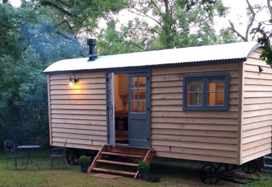 Het mobiele huis The Bakers Hut staat in het bos, met binnenverlichting en een kleine veranda aan de voorkant.