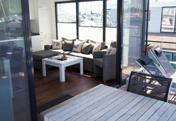 Sala de estar con sofá, mesa de centro y grandes ventanas en la casa flotante «Familiy & Friends LP 1 / 4 Personen» en Alemania.