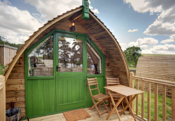 Een houten pod met groene deuren, terras, twee stoelen en tafel, in een landelijke, groene omgeving.