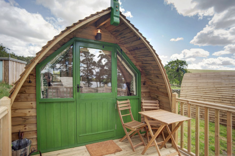 Een houten pod met groene deuren, klein terras, twee stoelen en een tafel, omringd door natuur.
