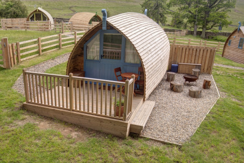 Foto van een gezellige houten pod met veranda, buitenterras en vuurplaats in een groene landelijke omgeving.