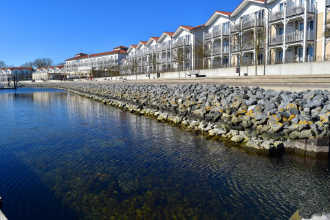 View from Houseboat Family & Friends LP 3 of calm water, stone shore, and modern apartments in Germany.