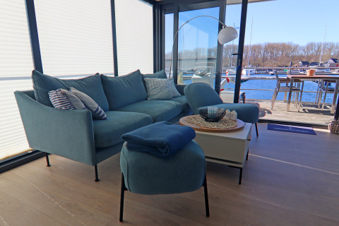 Lyst og hyggeligt stueområde på en husbåd med blå sofa, udsigt over vandet og udendørs spiseplads.