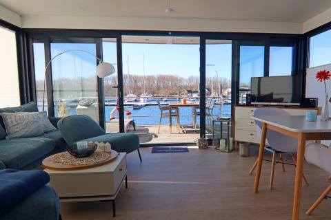 Lyst stueinteriør på en husbåd med udsigt til terrasse og marina, bådliv i baggrunden i Tyskland.