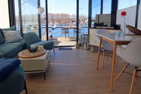 Sala de estar de una casa flotante con vistas al puerto deportivo, muebles modernos, sofá y mesa de comedor.