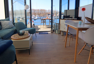 Wohnbereich eines Hausboots mit Blick auf den Hafen, modernen Möbeln, Sofa, TV und Esstisch.