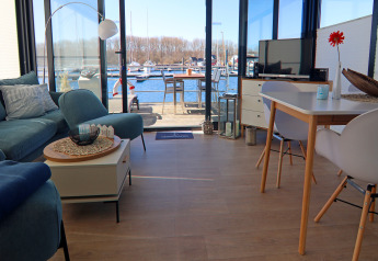 Wohnbereich eines Hausboots mit Blick auf den Hafen, modernen Möbeln, Sofa, TV und Esstisch.