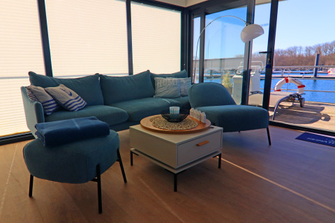 Wohnzimmer auf einem Hausboot mit blauer Couch, Tisch und großem Fensterblick auf Marina in Deutschland.