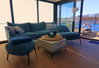 Living room on a houseboat with blue sofa, coffee table, and large windows overlooking a marina in Germany.