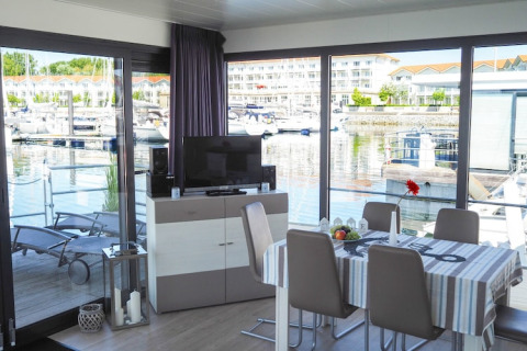 Interior de una casa flotante con mesa de comedor, televisor y grandes ventanas que dan al puerto deportivo.