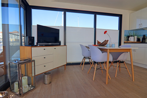 Modern living area on a houseboat with dining table, four chairs, TV unit, and large windows view.