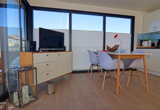 Modern living area on a houseboat with dining table, four chairs, TV unit, and large windows view.