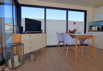 Moderno salón en una casa flotante con mesa de comedor, cuatro sillas, TV y grandes ventanales.