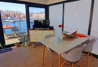Moderne spiseplads og tv-område på husbåd med udsigt til marinaen gennem store vinduer i Tyskland.