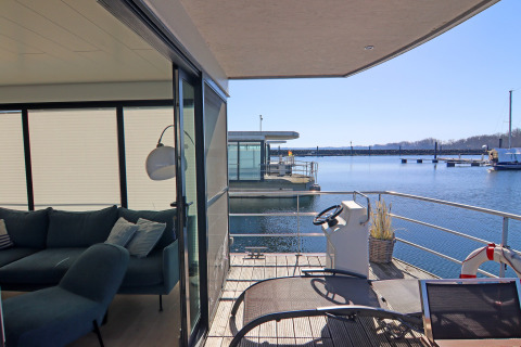 Udsigt fra en husbåd i Tyskland med terrasse, styrekonsol, loungestol og sofa i stuen.