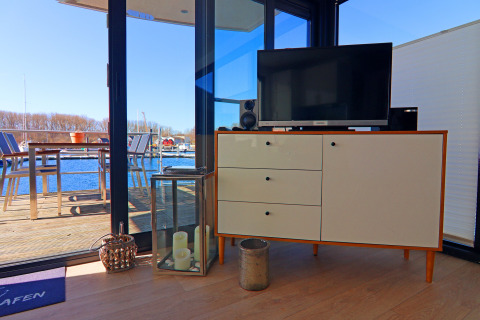 Wohnbereich eines Hausbootes mit Kommode, Fernseher, Kerzen und Ausblick auf Wasserterrasse und Yachthafen.