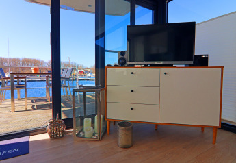 Salon d’une péniche avec buffet, télévision, bougies et vue sur la terrasse et le port par de grandes baies.