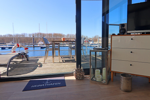 Udsigt fra en husbåd over en terrasse med bord og stole, med marina og både i baggrunden på en solrig dag.