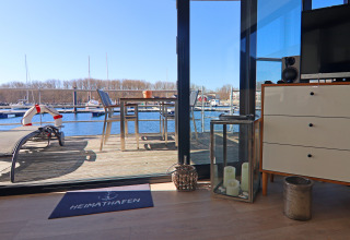 Vista desde una casa flotante hacia terraza con mesa y sillas, frente a un muelle deportivo y veleros al fondo.