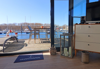 View from a houseboat through glass doors to a deck with marina, boats, a table and chairs on a sunny day.