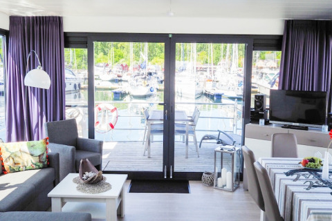 Moderne stue på husbåd med udsigt til marina, både og terrasse gennem store glasdøre, Tyskland.