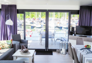 Salón moderno de casa flotante con vista al puerto deportivo, barcos y terraza a través de puertas de cristal en Alemania.