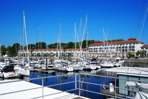 Casa flotante y veleros amarrados en un puerto deportivo con edificios blancos y cielo azul, Alemania.