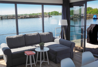 Wohnzimmer mit grauem Sofa, Tisch und Seeblick auf Hausboot Family & Friends mit Hund in Deutschland.