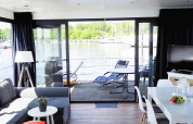 Indretning af en husbåd med udsigt over vandet, moderne stue og spisebord, udgang til terrasse.