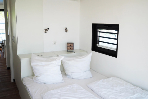 Dormitorio moderno en la casa flotante Family & Friends con perro, ropa de cama blanca y ventana pequeña.