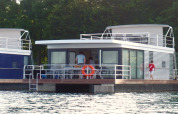 Moderne woonboot met glazen ramen op het water, personen zichtbaar op het dek, omringd door bomen.