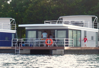 Moderne woonboot met glazen ramen op het water, personen zichtbaar op het dek, omringd door bomen.