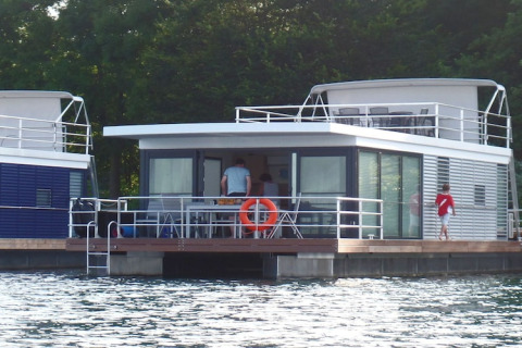 Moderne woonboot met glazen ramen op het water, personen zichtbaar op het dek, omringd door bomen.