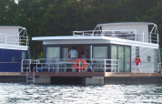 Houseboat moderne à grandes fenêtres sur l'eau, personnes visibles sur le pont, arbres en arrière-plan.
