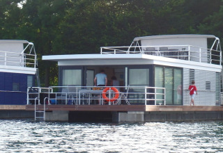 Houseboat moderna con grandi finestre sull’acqua, persone sul ponte, circondata da alberi verdi.