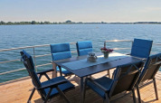 Eettafel met zes blauwe stoelen op terras van woonboot, uitzicht over kalm water en een heldere hemel.