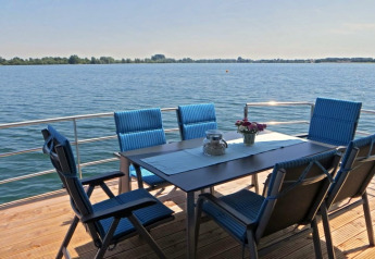Eettafel met zes blauwe stoelen op terras van woonboot, uitzicht over kalm water en een heldere hemel.