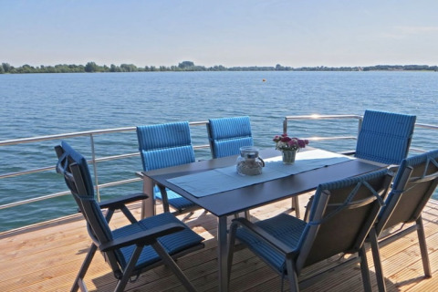 Eettafel met zes blauwe stoelen op terras van woonboot, uitzicht over kalm water en een heldere hemel.