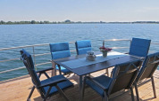 Table de jardin avec six chaises bleues sur la terrasse d'une péniche, vue sur un lac paisible et le soleil.