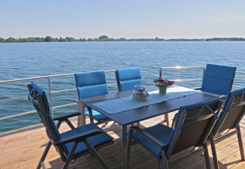 Table de jardin avec six chaises bleues sur la terrasse d'une péniche, vue sur un lac paisible et le soleil.