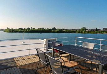 Terras van een woonboot met metalen tafel en stoelen, uitzicht op een rustige plas en groene natuur.
