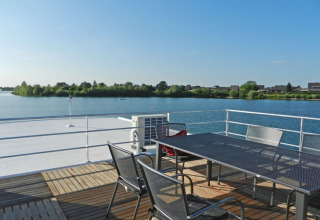 Terras van een woonboot met metalen tafel en stoelen, uitkijkend over een rustig meer en groene bomen.