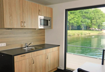 Moderne woonbootkeuken met houten kasten en groot raam met zicht op het meer en groene bomen.