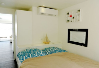 Slaapkamer op een woonboot voorzien van dubbel bed, airco, modelzeilboot en klein raam met rolgordijn.