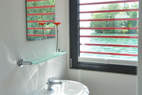 Bagno di una houseboat con finestra vista acqua, mensola di vetro, specchio e due fiori arancioni.
