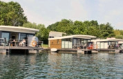 Modern houseboats are moored on a calm lake, surrounded by lush green trees and a clear blue sky.